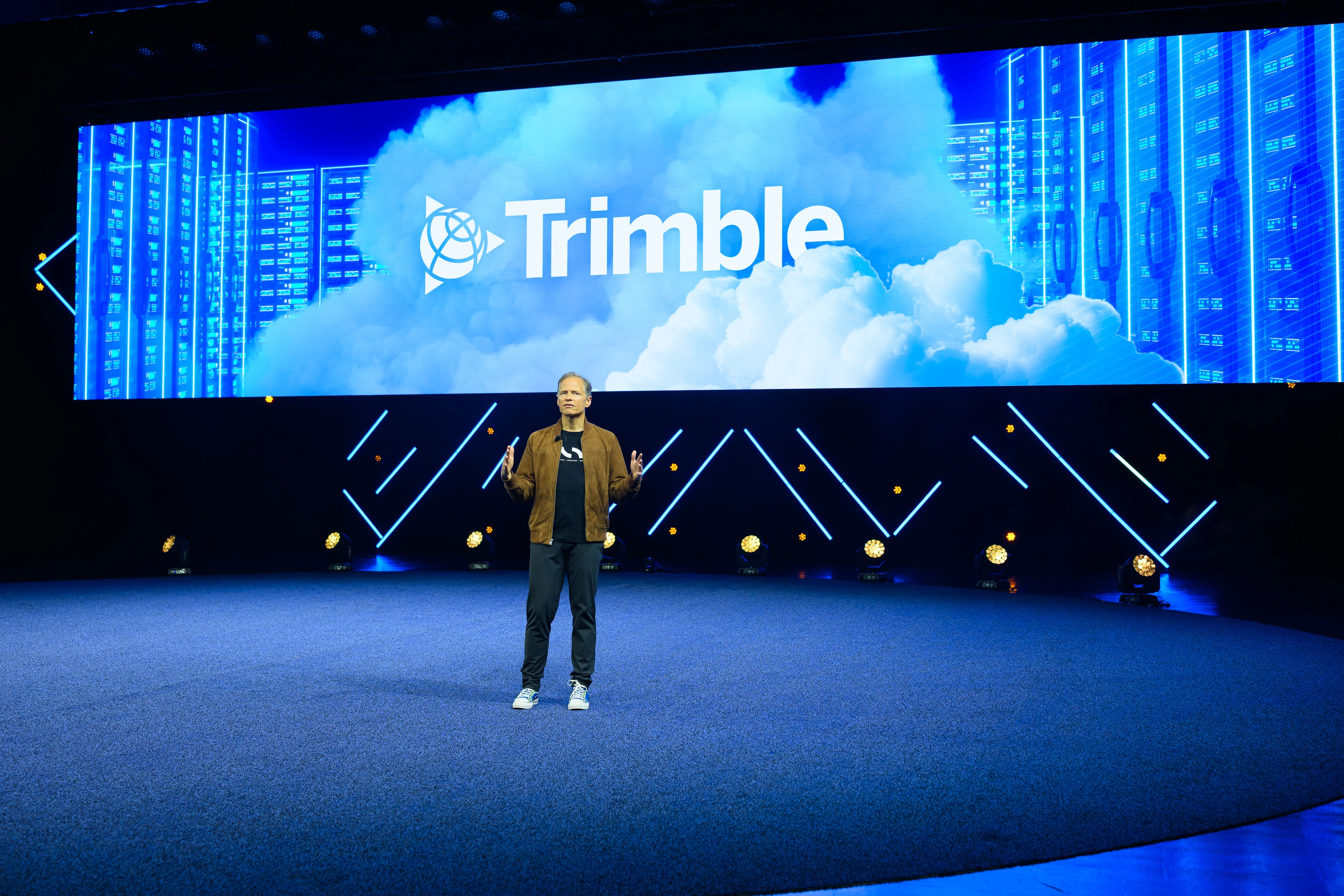 Keynote speaker on stage before a large Trimble logo and digital server backdrop at the Trimble Insight transportation technology conference.